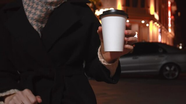 Two disposable cups made of white paper on blurred background of evening city lights. Woman taking one cup of coffee from parapet and drinking. Hot Takeaway Coffee Outdoors. Street food,