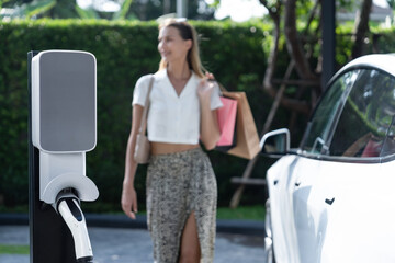 Young woman travel with EV electric car charging in green sustainable city outdoor garden in...