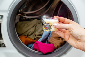 Person adding laundry detergent pod to the washer full with clothes. Female hand holding laundry...