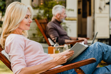 Content senior blond lady reading a book in the foreground with her grey-haired husband on the background in the garden with their trailer behind