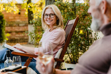 Smiling beautiful mature lady in glasses with glass of wine sitting in deck chair and talking with...