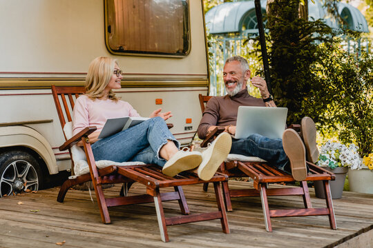 Senior caucasian cheerful couple talking in the camper van yard sitting on deck chairs with book and laptop, traveling together, caravanning by trailer motor home
