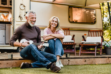 Senior caucasian man husband playing ukulele and his blonde attractive wife listening to his play in the camper van trailer motor home porch. Togetherness