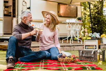 Joyful senior couple spouses travelers husband and wife toasting with glasses of champagne celebrating sitting on the blanket in the caravan porch, on a trip together in camper van
