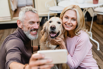 Nice-looking senior caucasian couple taking selfie with their dog on the caravan porch. Vlogging blogging online while traveling with golden retriever by trailer motor home