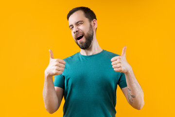 Studio shot of cheerful confident young man posing over yellow background winking and holding thumbs up - obrazy, fototapety, plakaty