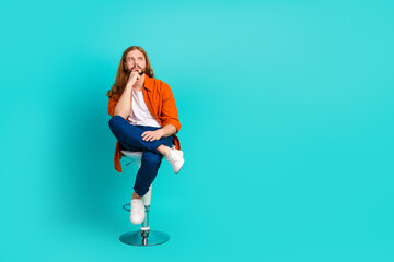 Fototapeta na wymiar Full length photo of clever man dressed orange shirt sit on chair hand on chin look at offer empty space isolated on teal color background
