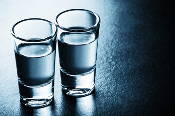 Glass vodka shots. Two vodka glass. Weekend party background. Blue bar surface reflection. Backlight transparent glass backdrop. Color vodka background. Couple drinking. Empty copy space on the right.