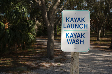 Kayack Launch sign white and blue on a wood post with trail and green trees in the background....