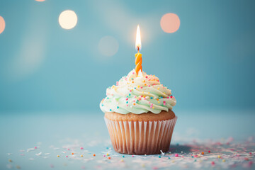 Single cupcake with frosting, sugar sprinkles and burning birthday candle in front of blue background