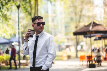 Professional, modern and confident young businessman with white shirt and black tie holding a eco-friendly paper cup of coffee in urban park of business district