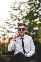 Professional, modern and confident young businessman with white shirt and black tie making a call with a light blue smartphone in urban park of business district