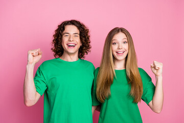Photo of two optimistic teenagers friends wearing green t shirt raised fists up winning lottery together isolated on pink color background