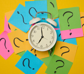 A round alarm clock amidst paper stickers featuring drawn question marks, symbolizing the concepts...