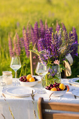 Romantic table decor for a loving couple on the blooming meadow with purple lupines. Two glasses of wine, flowers in a vase, silverware, fruits, wooden furniture and picnic basket. Sunset, golden hour