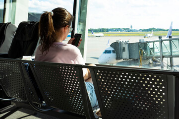 Young woman waiting for departure while using smart phone in airport lounge. Female traveler and tourist theme. High season and vacation concept. Airplane background.