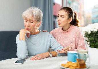 Apologetic adult daughter calming her upset elderly mother at table at home, apologizing after dispute. - obrazy, fototapety, plakaty