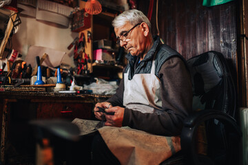 An old craftsman is sitting at his workshop and cutting leather parts.