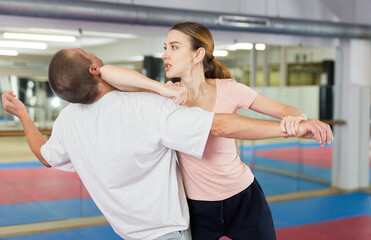 Fototapeta na wymiar Concentrated sporty young woman learning self defence techniques in sparring with man, practicing elbow blow with wristlock to opponent in gym