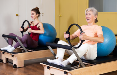 Cheerful active elderly woman exercising with flex ring and fitball on reformer in pilates studio during group workout