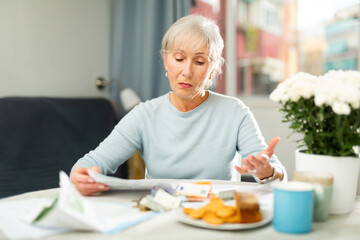 Portrait of troubled elderly woman looking at papers at home, worrying about debts on bills.