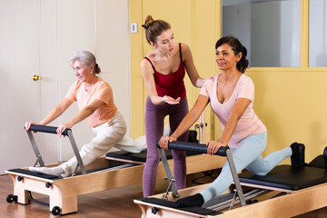 Two health conscious women of different ages perform a Pilates exercise using a reformer bed, where a female instructor ..helps them do it correctly