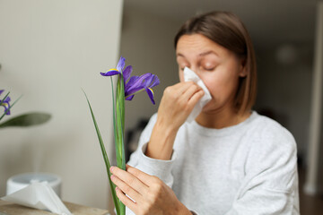 Allergic young woman holds iris flower, covers nose with paper tissue has runny nose, sneezes from flowers pollen at kitchen home by table. Girl with flu, itching or cough, seasonal allergy, rhinitis