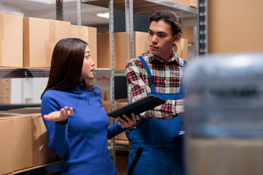 Warehouse asian workers discussing stock tracking while managing product inventory in storage room. Storehouse employees doing financial management on digital tablet and talking near boxes shelf