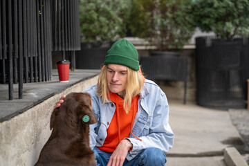 Caring young guy in stylish clothes strokes a homeless microchipped dog on the street on summer day in the park. Concept of caring for abandoned animals. Volunteer man at a shelter for homeless pets.