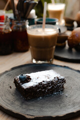 Chocolate brownie dipped in powdered sugar with a blueberry, in the background a glass of coffee, sauces and more food
