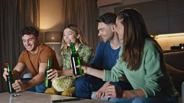 Chilling friends clinking beer bottles relaxing at home couch. Smiling mates