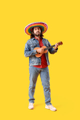 Handsome young Mexican man in sombrero playing guitar on yellow background