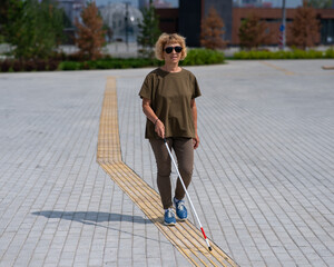 An elderly blind woman walks with a cane along a tactile tile. 