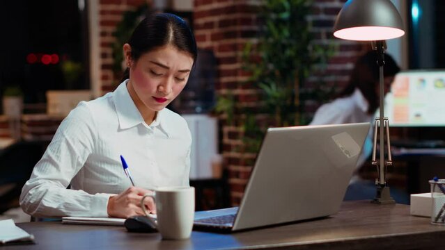 Asian worker sulking in office, having frown on face, feeling annoyed by having too much work to do. Employee writing down info on notepad, gloomily solving tasks late at night