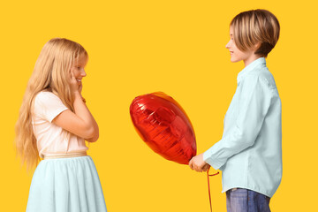 Little boy giving heart-shaped balloon to girl on yellow background. Valentine's Day celebration