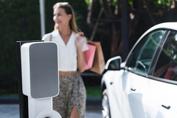 Young woman travel with EV electric car charging in green sustainable city outdoor garden in...