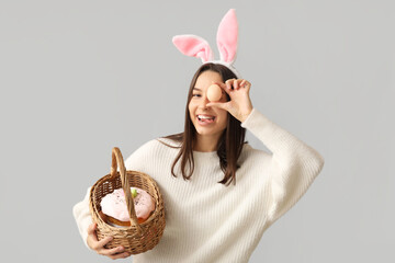 Pretty young woman with bunny ears and Easter basket on grey background