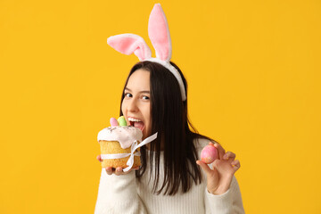 Pretty young woman with bunny ears, Easter cake and egg on yellow background