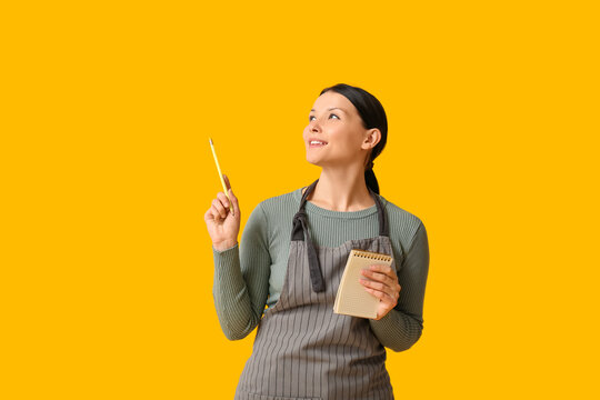 Beautiful young waitress with notebook and pencil on yellow background