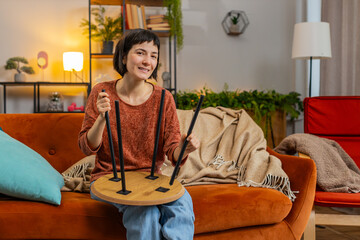 Happy smiling Caucasian woman finishing assembling furniture at home sitting on orange couch in living room. Successful table collect, repair, fixing. Girl after moving into new apartment. Mortgage.