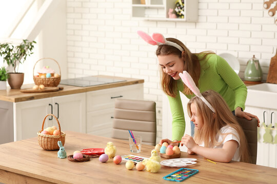 Cute little girl and her mother in bunny ears painting Easter eggs in kitchen