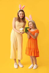 Cute little girl with rabbit and her mother in bunny ears holding Easter cake on yellow background