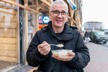 Mature solo traveler smiling and eating a street food e.g. rice. Man enjoying outdoor lifestyle...