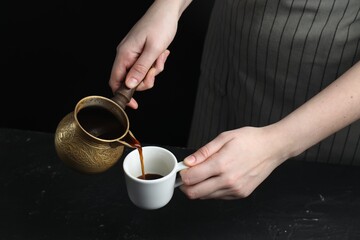 Turkish coffee. Woman pouring brewed beverage from cezve into cup at black table, closeup