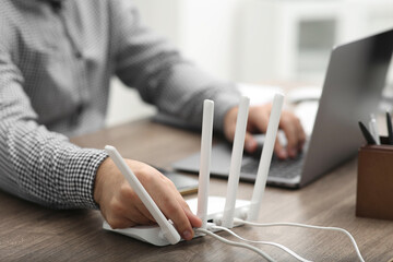 Man with laptop inserting cable into Wi-Fi router at wooden table indoors, closeup