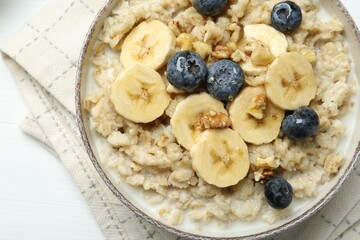 Tasty oatmeal with banana, blueberries, walnuts and honey served in bowl on white wooden table, top view
