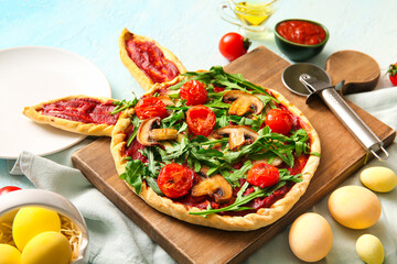 Wooden board with tasty pizza and painted eggs for Easter celebration on blue background
