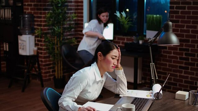 Bookkeeper feeling dread while having to do complicated tax reports in office at night. Worker squinting eyes in exasperation, feeling overworked while using laptop to do project research, camera A