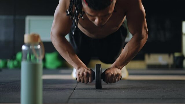 Close-up. A man with dreadlocks on his head pumps up his abs using a gymnastic roller indoors in a modern gym