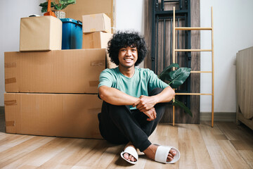 Cheerful young Asian male sitting on floor, looking at camera with happy face after moving in new...